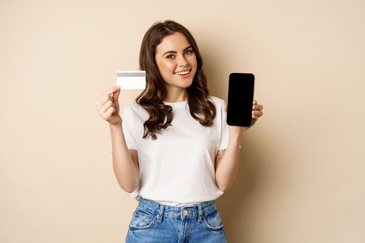 Online shopping and people concept. Young beautiful woman looking happy, showing credit card discount and mobile phone screen, standing over beige background.