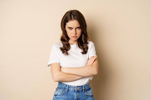 Offended angry woman cross hands on chest, frowning and sulking insulted, standing over beige background.