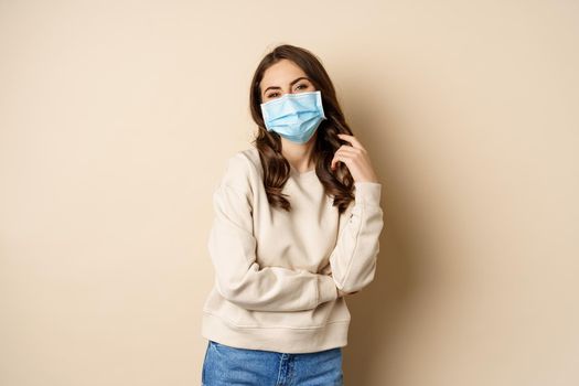 Covid-19, pandemic and quarantine concept. Beautiful modern woman in medical face mask, cross arms on chest, looking confident, beige background.