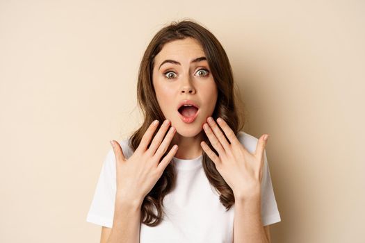 Close up portrait of surprised and excited brunette girl looking amazed, reacting impressed and excited, standing against beige background.