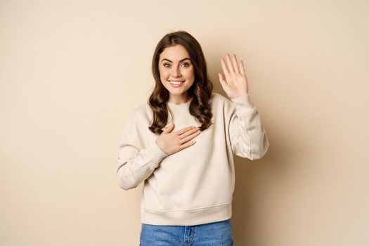 Cute modest girl smiling, raising finger, introduce herself, saying hello, standing over beige background.