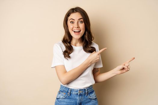 Smiling brunette woman in t-shirt, pointing fingers right, showing promo offer or advertisement, demonstrating banner, standing against beige background.