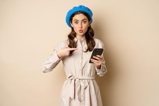 Excited girl with smartphone pointing finger at mobile screen, looking amazed, wow intrigued face expression, beige background.