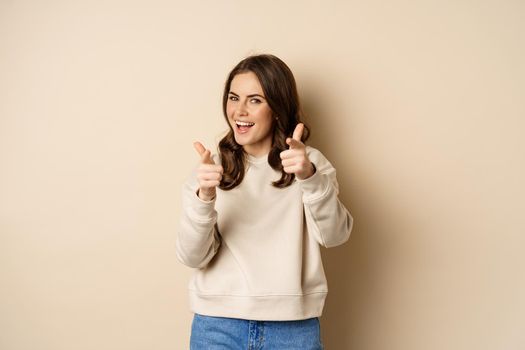 You got this. Happy young woman pointing fingers at camera, praising, complimenting, congrats gesture, standing over beige background.