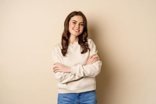 Beautiful young brunette woman posing happy against beige background, smiling at camera, wearing sweater.