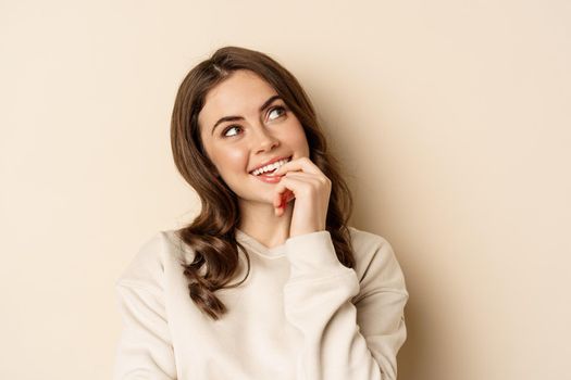 Cute modern woman thinking, daydreaming and smiling, looking up thoughtful, standing over beige background.