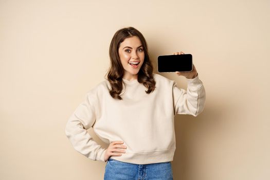 Smiling beautiful female model demonstrating horizontal smartphone screen, mobile application, standing over beige background.