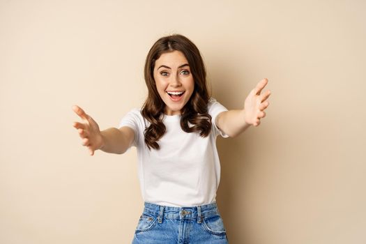 Portrait of happy young woman smiling, stretching arms out for hug, cuddling, reaching hands to hold receive smth, standing over beige background.