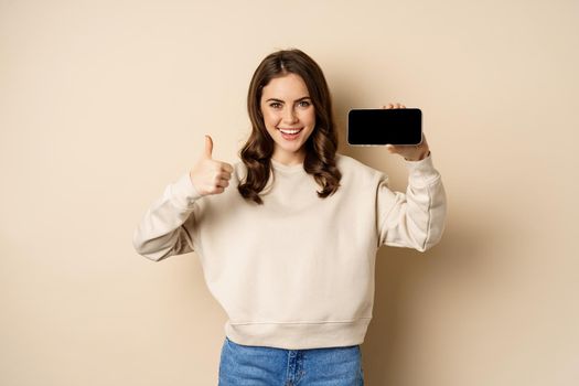 Smiling happy woman showing smartphone horizontal screen, thumbs up, recommending website, online store or app, standing over beige background.