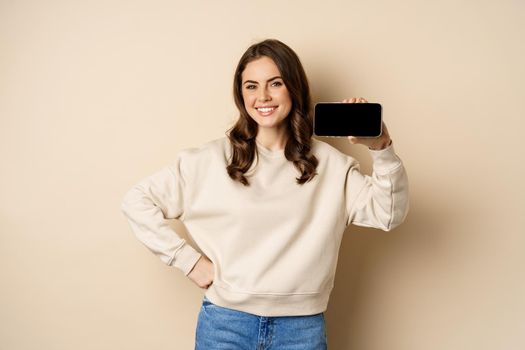 Portrait of smiling cute woman showing horizontal smartphone screen, website or application, standing over beige background.