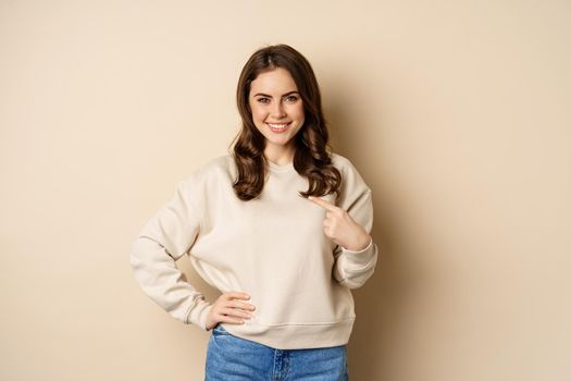 Confident smiling modern woman pointing finger at herself, self-promoting, standing over beige background. Copy space