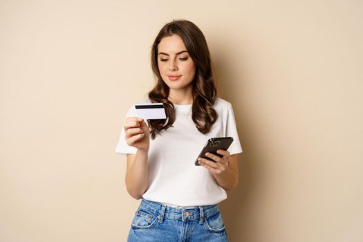 Image of young happy woman paying online, holding smartphone and credit card, enter info in application on mobile phone, standing against beige background.