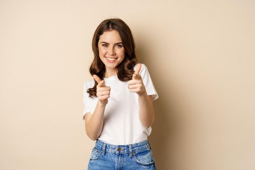 You got this, congrats. Smiling young woman pointing fingers at camera, cheering, complimenting, standing over beige background.
