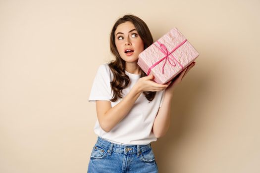 Celebration and holidays concept. Happy young girl looking intrigued, shaking box with gift, guessing whats inside wrapped present, standing over beige background.