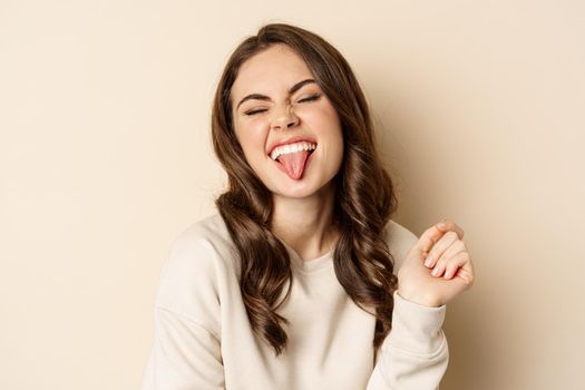 Close up of carefree happy woman, showing tongue and having fun, posing in sweater over beige background. Copy space