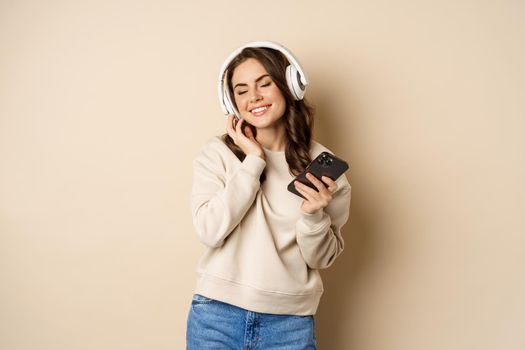 Happy caucasian woman listening music in headphones on smartphone app, dancing and having fun, standing over beige background.