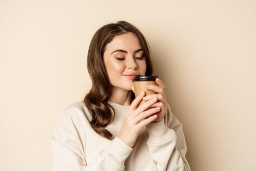 Woman smiling and smelling delicious cup of coffee in takeaway cup, standing over beige background.
