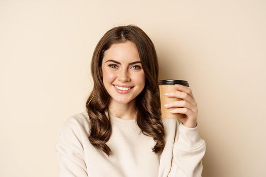 Takeaway and cafe concept. Beautiful feminine woman smiling, holding cup of coffee, posing against beige background. Copy space