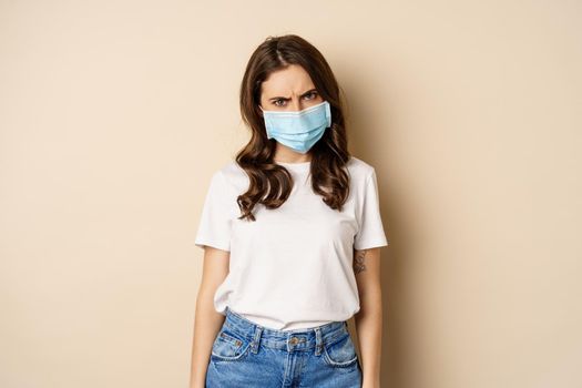 Image of angry girl in medical face mask, grimacing and looking insulted, standing displeased against beige background.