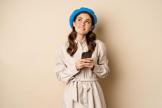 Dreamy smiling woman in stylish trenchcoat, looking up fantasizing while shopping on mobile phone app, using smartphone and thinking, standing over beige background.