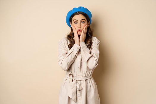 Image of stylsh beautiful woman looking surprised, shocked reaction at camera, posing in trench coat against beige background.