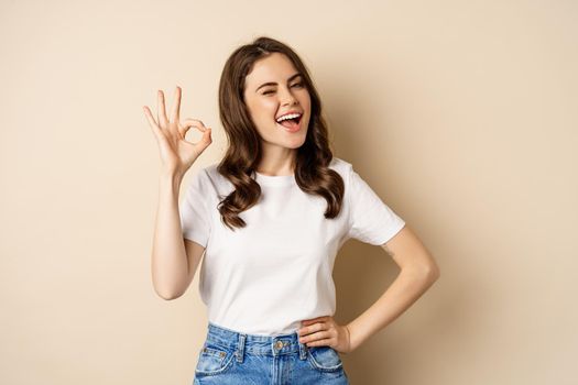 Stylish young woman smiling satisfied, showing okay, ok sign in approval, recommending, standing over beige background.