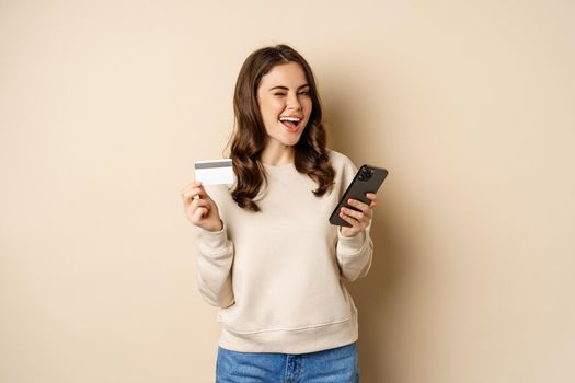 Smiling girl using mobile app, smartphone shopping and credit card, standing over beige background, order smth.
