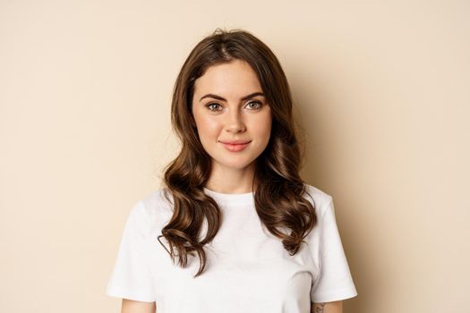 People and emotions. Close up of confident smiling young woman, looking at camera, standing in casual relaxed pose, wearing white t-shirt, beige studio background.