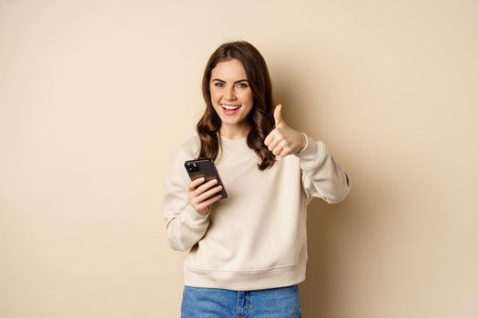 Young woman smiling, showing thumbs up while using mobile phone, smartphone app, standing over beige background.