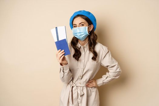 Covid pandemic and travelling concept. Young woman tourist showing her passport and tickets abroad, wearing face mask from coronavirus, posing over beige background.
