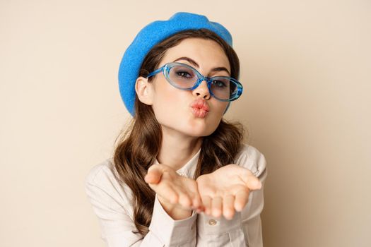 Beautiful stylish woman in sunglasses, blowing air kiss on hands near lips, gazing softly and flirty at camera, posing against beige background.