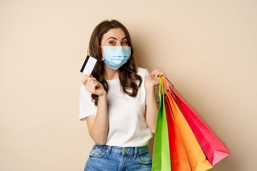 Covid-19 pandemic and lifestyle concept. Young woman posing in medical face mask with shopping bags from mall, holding credt card in hand, beige background.