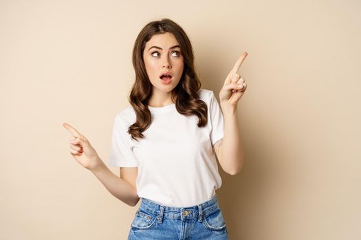 Happy authentic girl smiling, pointing fingers sideways, showing left and right banner, demonstrating promo, standing against beige background.