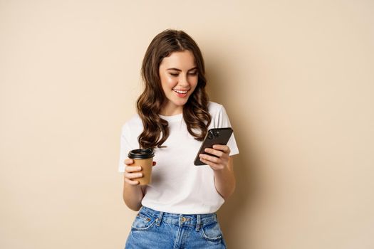 Smiling woman using mobile phone app and credit card, concept of online shopping, contactless payment and delivery, standing over beige background.