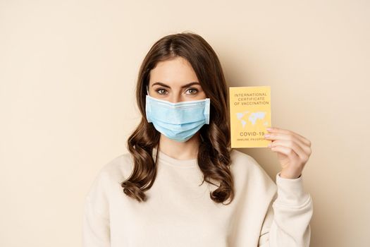 Covid and people concept. Vaccinated woman in face mask showing covid international vaccination certificate, recommend get vaccine, standing over beige background.