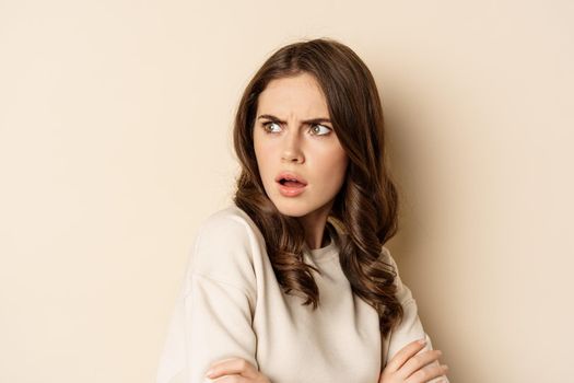 Close up of confused young woman, looking behind with skeptical shocked face expression, standing against beige background.