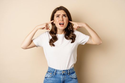 Annoyed young woman shut ears from loud noise, feeling discomfort and complaining, standing against beige background.