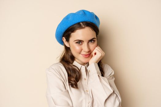 Close up portrait of feminine young woman in trendy french hat, posing cute and romantic, making silly coquettish face and tender smile, beige background.