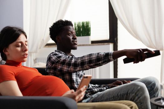 Relaxed young multiethnic couple sitting together on sofa while expecting newborn. Pregnant caucasian future mother using smartphone while african american man switching channels on TV utilizing remote.
