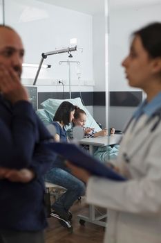 Recovering little daughter playing games on mobile phone while young mother sitting beside her bed. Worried father talking with pediatric specialist about consultation results and treatment recovery.