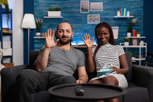 POV of interracial young couple waving at videoconference call while holding a bowl of popcorn. People talking to relatives through videocall communication technology while sitting in living room.