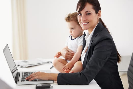 Young working mom smiling at the camera while holding a camera and typing on her laptop.
