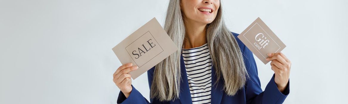 Cheerful middle aged Asian woman with hoary hair holds gift card and Sale sign standing on light grey background in studio, space for text