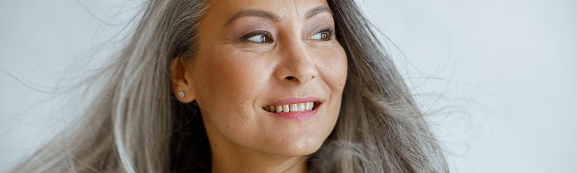 Happy middle aged Asian woman with flying grey locks stands on light background in studio closeup. Mature beauty lifestyle