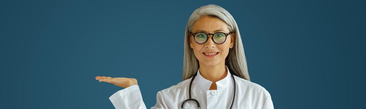 Positive middle aged lady doctor in white robe with glasses and stethoscope points aside standing on blue background in studio. Professional medical staff