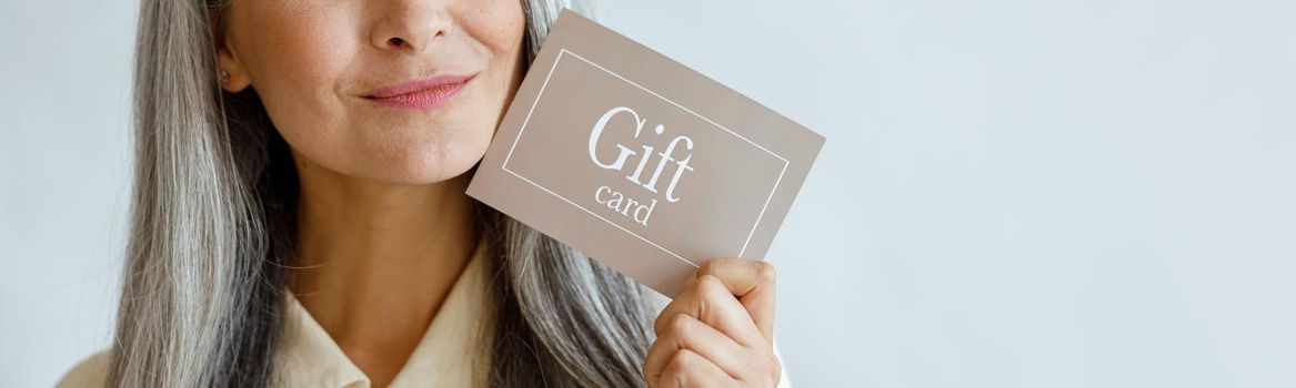 Dreamful grey haired Asian woman holds gift card near face standing on light grey background in studio, space for text. Shopping certificate