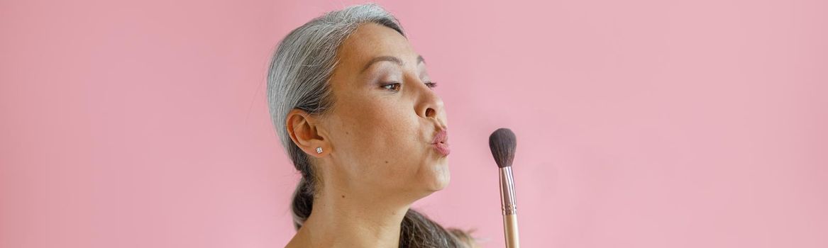Funny middle aged Asian lady wrapped with white towel blows cosmetic brush posing on pink background in studio. Mature beauty lifestyle