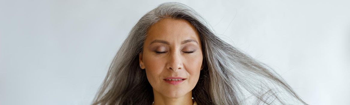Pretty tranquil Asian woman with flying silver hair stands on light grey background in studio. Mature beauty lifestyle