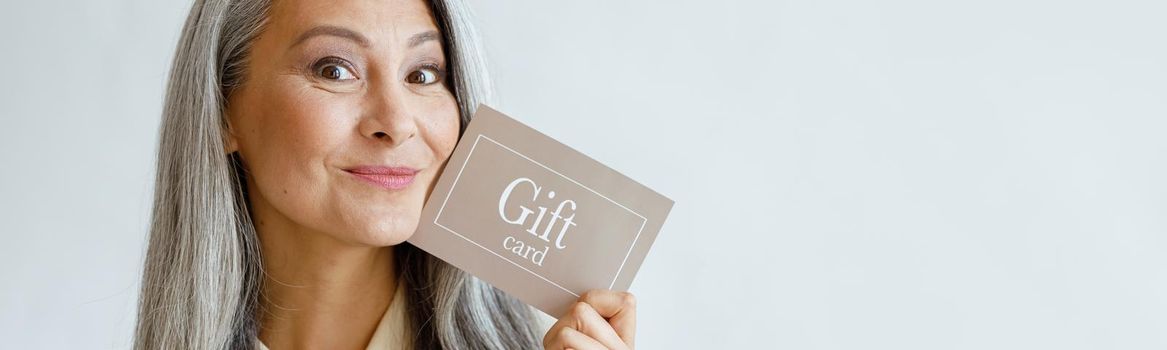 Joyful grey haired Asian woman holds gift card near face posing on light grey background in studio, space for text. Shopping certificate