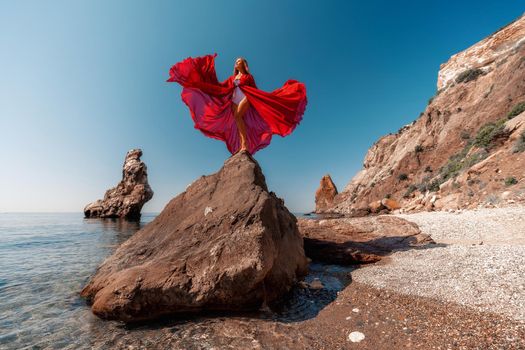 Beautiful woman posing in a luxurious red dress for the beach and in a white swimsuit with a long train, standing on a cliff against the sea. Around the cliff, the seashore